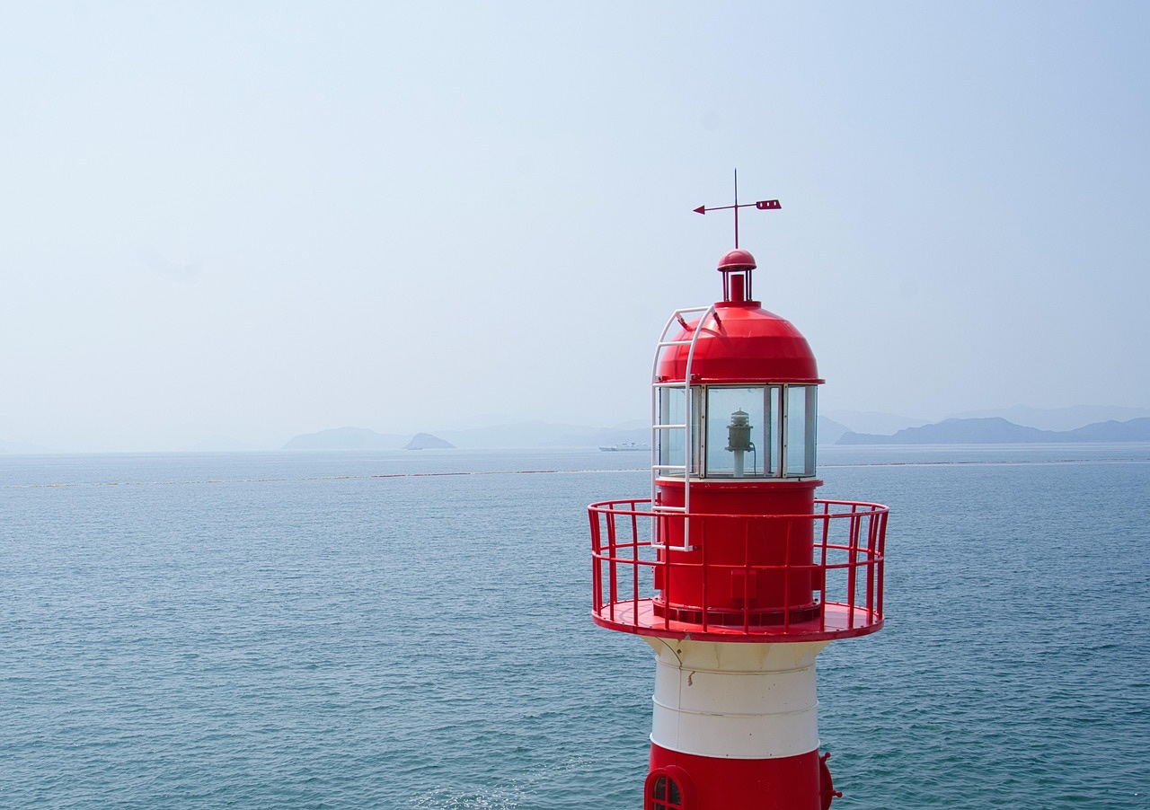 穏やかな青い海と遠くの島影を背に、赤と白の灯台が静かに立つ、よく晴れた日の海辺の風景写真